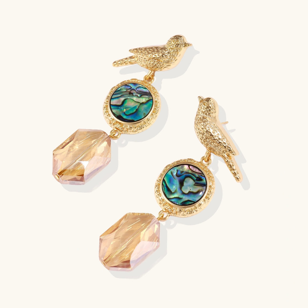 Gold-plated bird stud earrings with iridescent abalone shell medallions and champagne crystal drops on cream background
